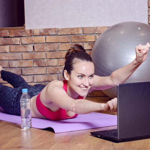 Young smiling woman lying on fitness Mat depicts the flight of a superhero during the warm-up before training online with videos on the laptop. funny home fitness.